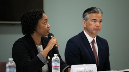 Image of Assemblymember Akilah Weber speaking at microphone. Attorney General Rob Bonta is in the background.