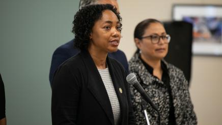 Image of Assemblymember Akilah Weber speaking at microphone. Norma Chavez-Peterson is in the background.