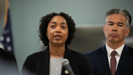 Image of Assemblymember Akilah Weber speaking at microphone. Attorney General Rob Bonta is in the background.